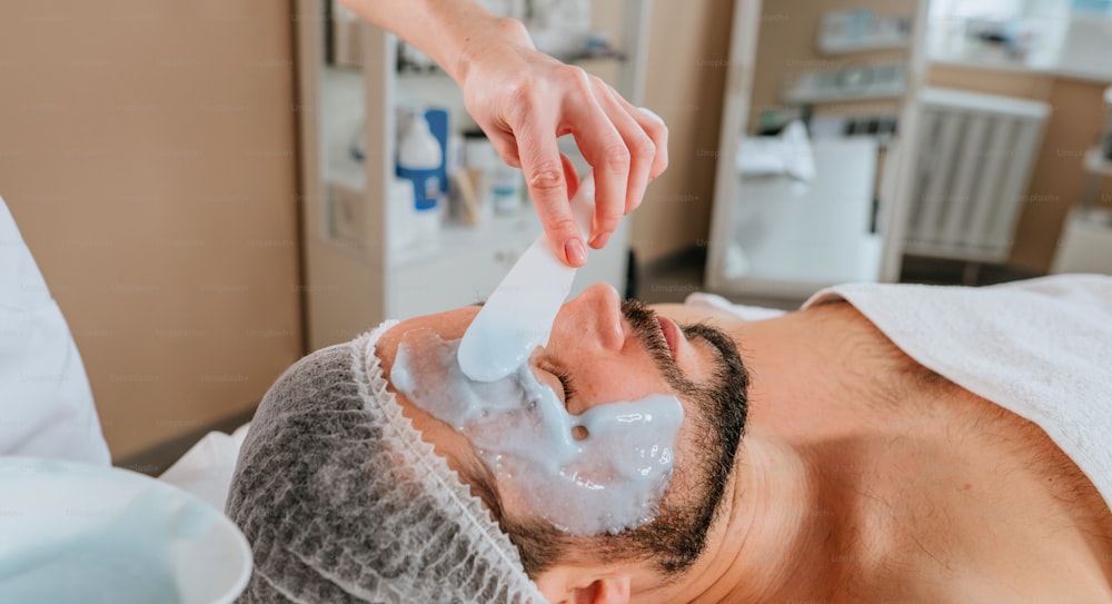 Esthetician making facial cleansing procedure using algae mask for a man in the beauty salon