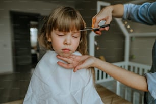 Nahaufnahme der Hände einer Mutter, die Haare ihrer vierjährigen Tochter zu Hause im Kinderzimmer schneidet. Heimalltag.