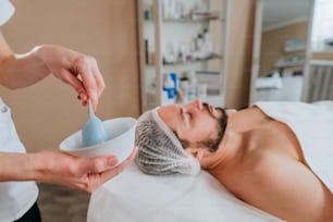 Esthetician making facial cleansing procedure using algae mask for a man in the beauty salon