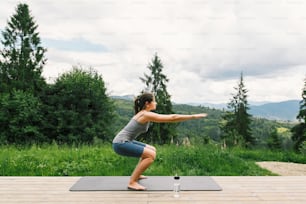 Jeune femme en forme s’étirant ou faisant des squats sur un tapis de yoga sur fond de collines de montagnes ensoleillées. Entraînement et entraînement en plein air. Femme décontractée sur une terrasse en bois parmi les arbres, mode de vie sain