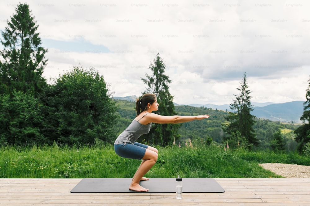 Young fit woman stretching or doing squats on yoga mat on background of sunny mountains hills. Outdoor workout and training. Casual female on wooden terrace among trees, healthy lifestyle