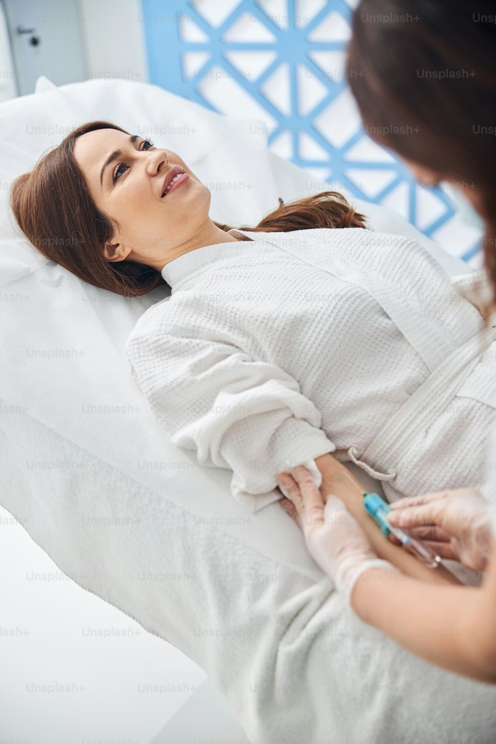 Charming lady in bathrobe lying on daybed and smiling while doctor making injection in her arm