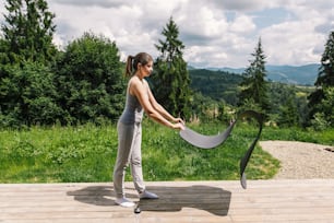 Young fit woman holding yoga mat for exercise on background of sunny mountains hills. Outdoor workout and training. Female in grey sport suit standing on wooden terrace among trees