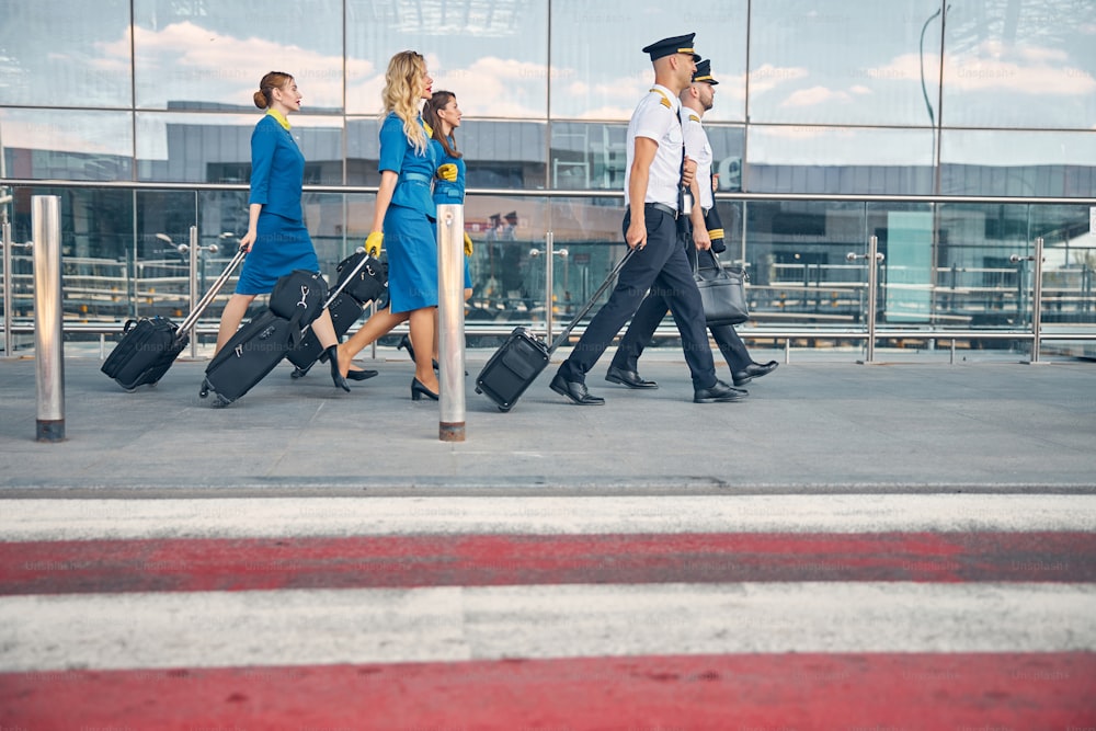 Hermosas azafatas y guapos pilotos con bolsas de equipaje con carro que se pasean por la carretera en el aeropuerto