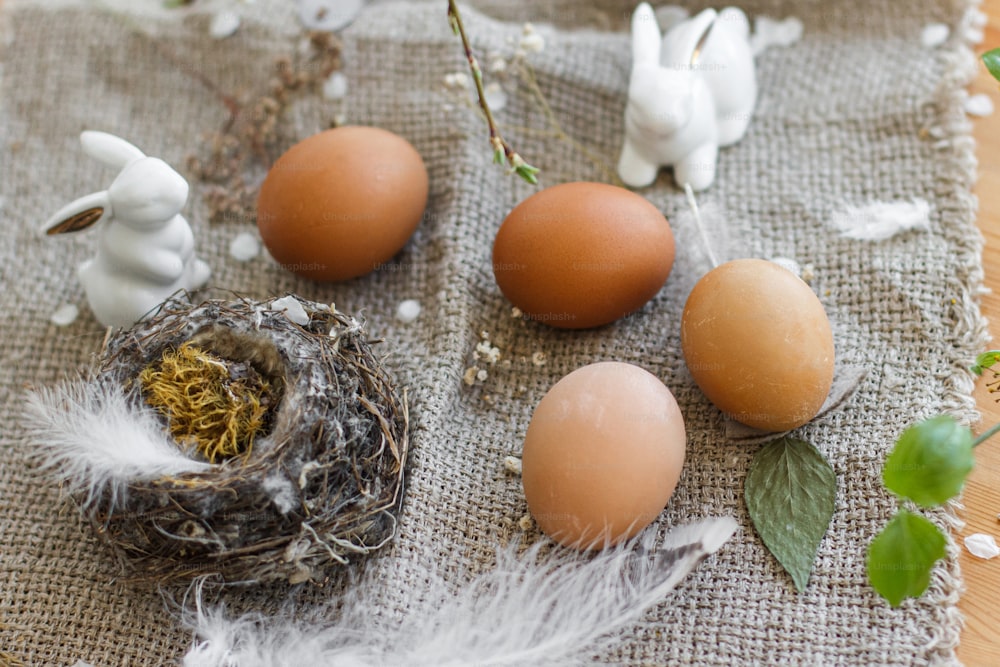 Huevos de Pascua naturales, conejitos blancos, plumas, nido y rama de cerezo en flor con pétalos en servilleta de lino rústico sobre mesa. Felices Pascuas. Espacio para el texto. Decoración respetuosa con el medio ambiente