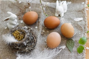 Ovos de Páscoa naturais, coelhos brancos, penas, ninho e ramo florido de cereja com pétalas em guardanapo de linho rústico sobre a mesa. Feliz Páscoa. Espaço para texto. Decoração ecológica