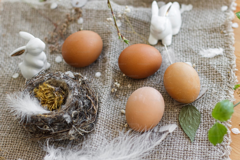 Natural Easter eggs, white bunnies, feathers, nest and cherry blooming branch with petals on rustic linen napkin on table. Happy Easter. Space for text. Eco friendly decor