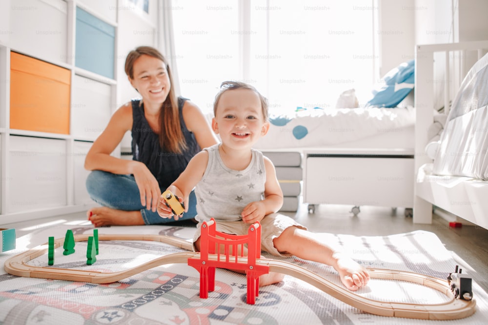 Mother and toddler boy playing with car wooden railway on floor at home. Early age education development. Kids building rail road and playing educational toy trains cars. Leisure activity for kid.