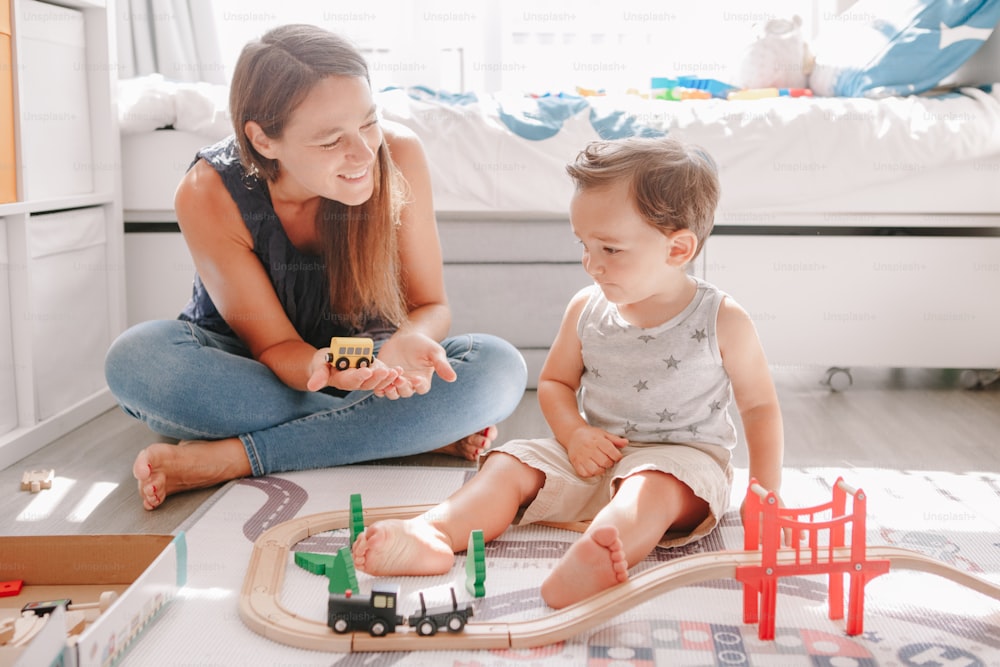 自宅の床で木造の鉄道で遊んでいる母親と幼児の男の子。幼児教育の発達。線路を建設したり、教育用のおもちゃの電車で遊んだりする子供たち。子供のための余暇活動。