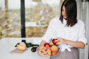 Young happy woman holding bowl with summer fruits at white countertop with vegetables in modern white kitchen. Healthy eating and Home cooking concept.