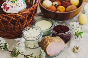 Delicious Easter food, stylish easter eggs, beets, cheese, butter, ham, homemade Easter bread and wicker basket with blooming spring flowers on linen napkin on rustic table. Happy Easter!