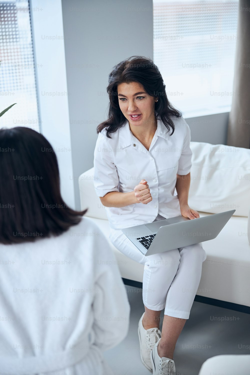 Une travailleuse de la santé concentrée avec un ordinateur portable sur les genoux interviewant une femme pendant la consultation