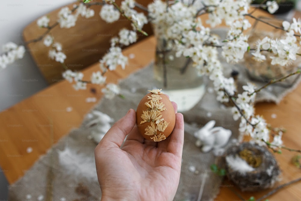 Hand hält Osterei verziert mit trockenen Blütenblättern auf dem Hintergrund des rustikalen Tisches mit Leinenserviette, Kirschblüte und Hase. Kreatives natürliches, umweltfreundliches Dekor von Ostereiern. Frohe Ostern