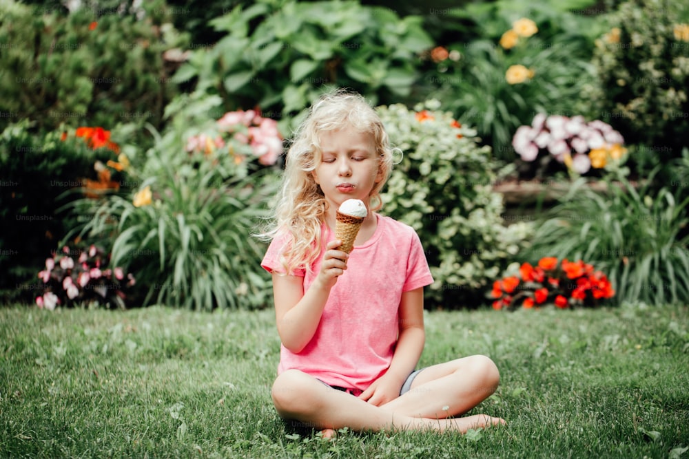 Ragazza adorabile divertente carina seduta sull'erba nel parco che mangia leccando il gelato dal cono di cialda. Bambino che mangia gustoso cibo estivo dolce freddo all'aperto. Spuntino estivo surgelato.