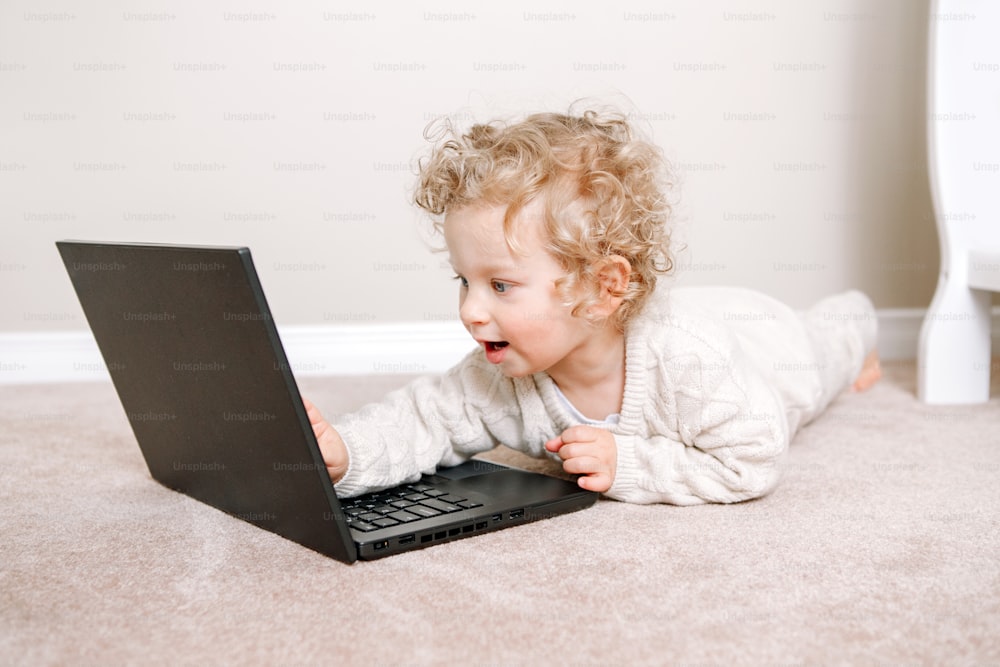 Bonito loiro encaracolado bebê bebê trabalhando no laptop. Criança pequena usando a tecnologia. Desenvolvimento da educação em idade precoce. Chat por vídeo, chamada de vídeo. Tempo de tela do computador para crianças.