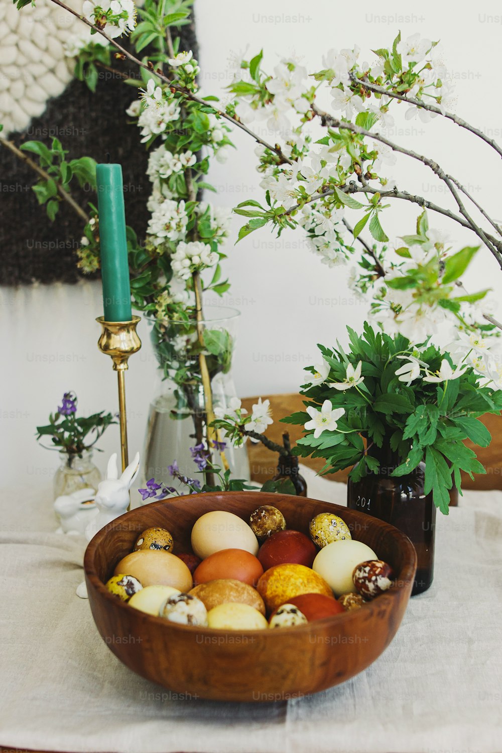Elegantes huevos de pascua en cuenco de madera sobre mesa rústica con figuritas de conejitos, velas y flores que florecen en primavera. Huevos teñidos al natural en colores amarillo y rojo. ¡Felices Pascuas!