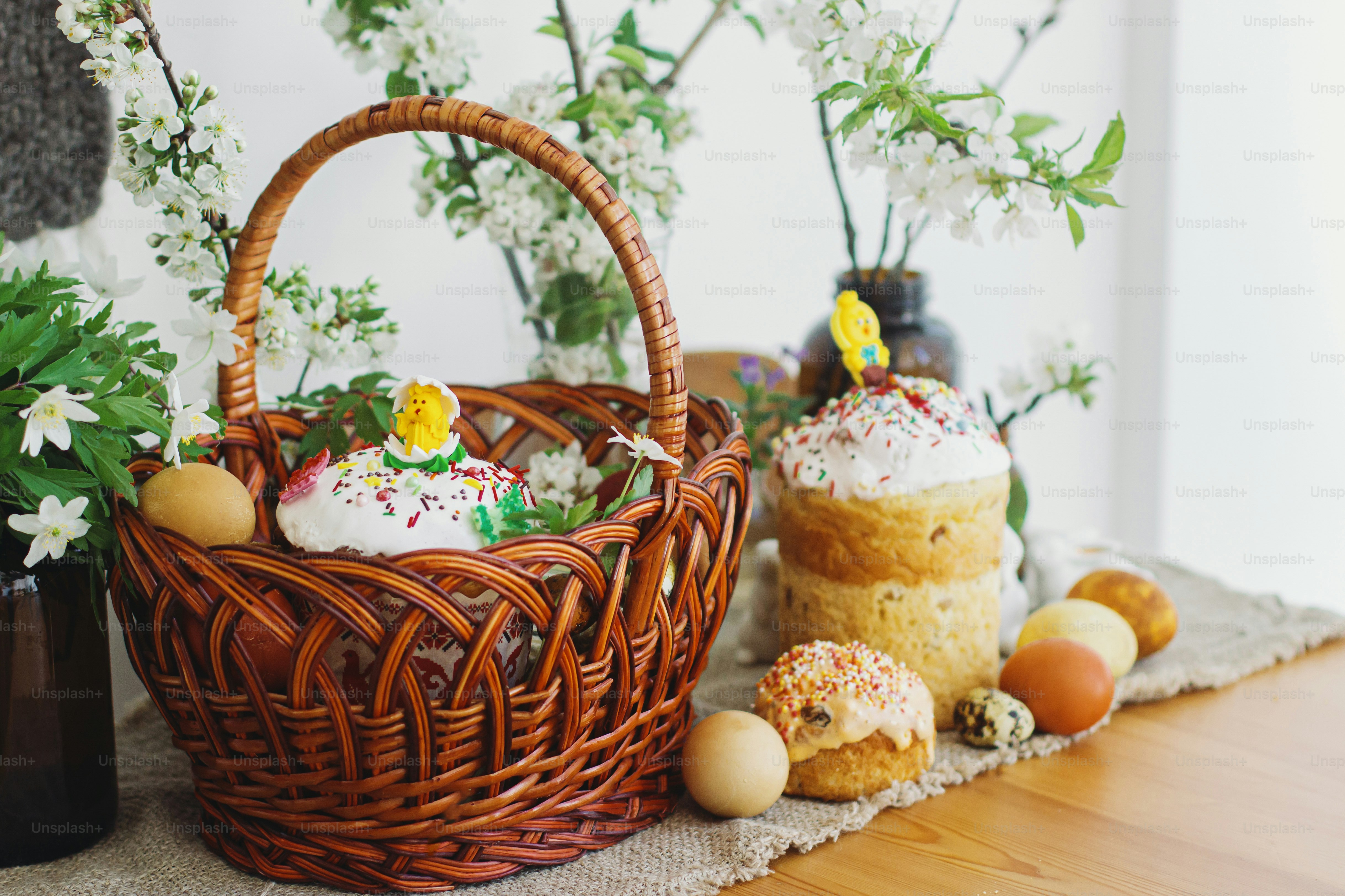 Stylish easter eggs, homemade Easter bread, delicious traditional Easter food in wicker basket and blooming spring flowers on linen napkin on rustic table. Happy Easter! Festive breakfast