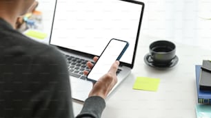 Vue par-dessus l’épaule d’un jeune homme indépendant utilisant un téléphone portable et travaillant avec un ordinateur portable sur un lieu de travail moderne.