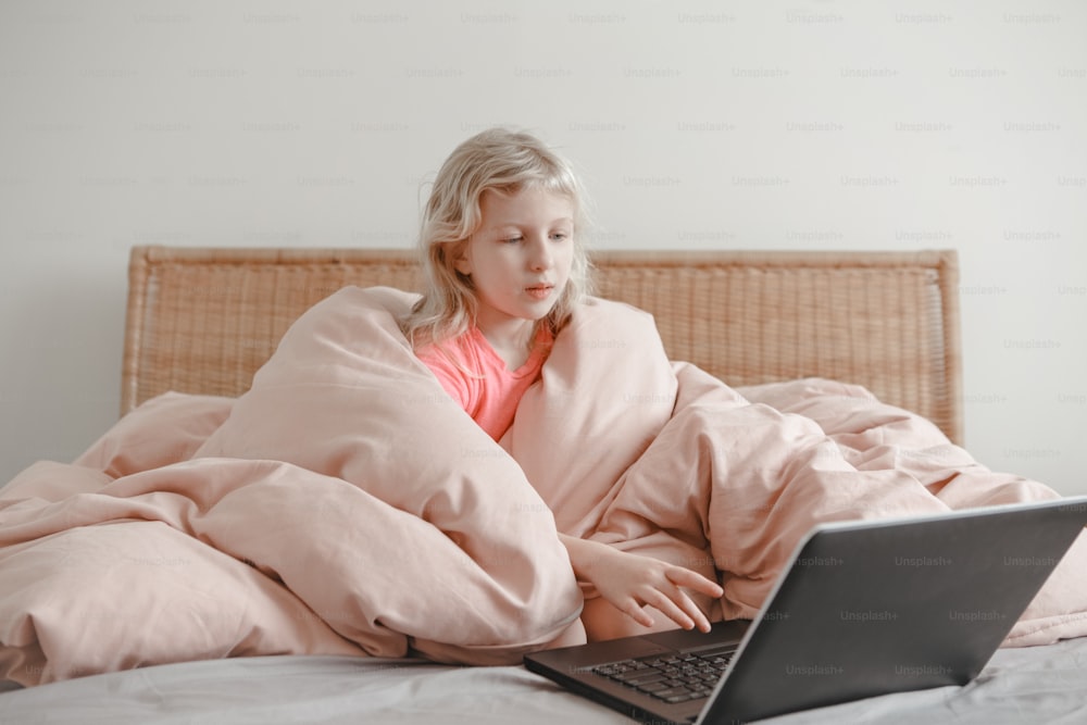 Girl sitting in bed under blanket and learning in virtual online school class. Kid working on laptop Internet at home. Child using wireless technology education. Homeschooling for children.