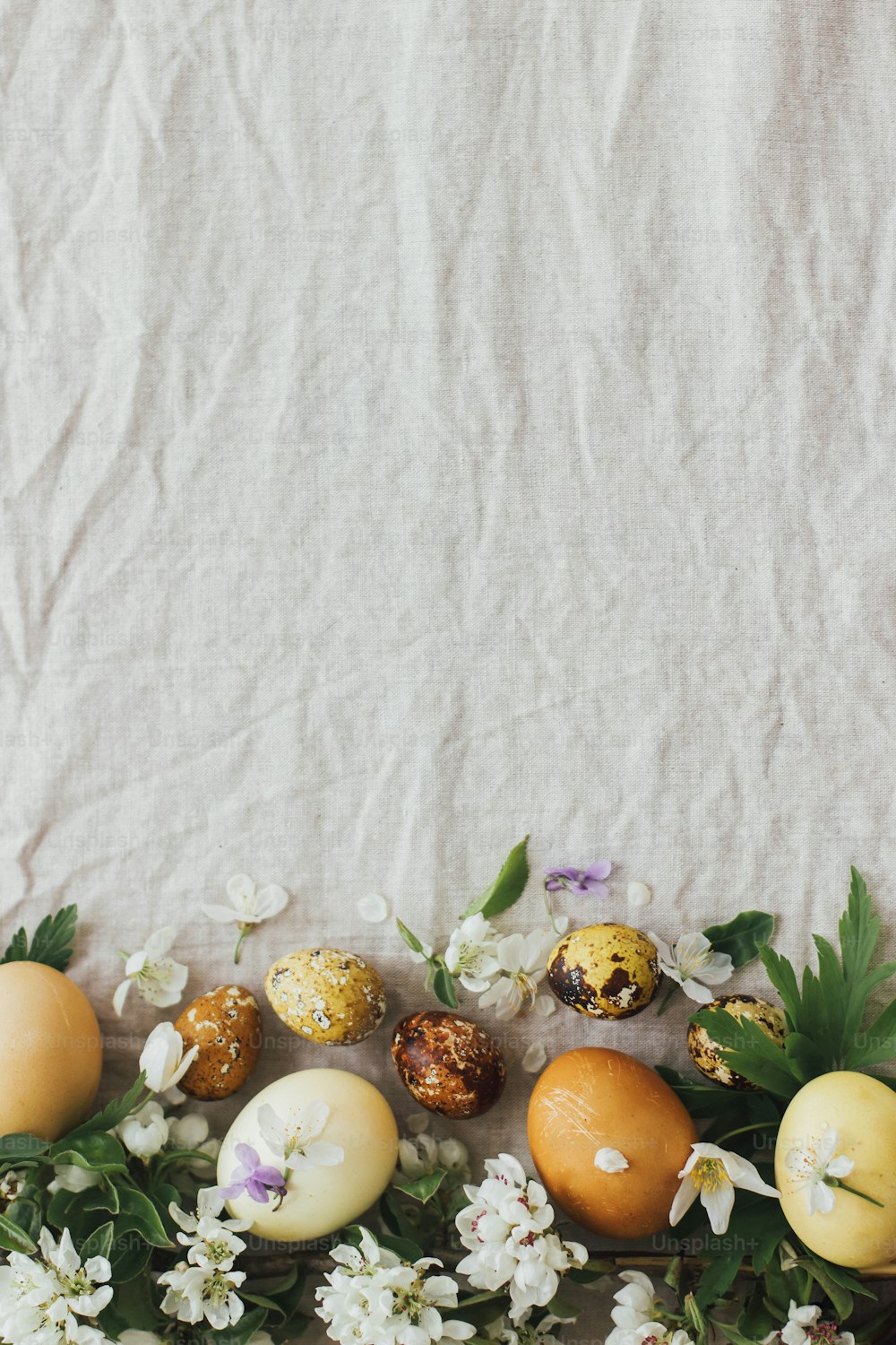 Easter eggs with spring flowers on rustic linen background, flat lay with space for text. Stylish modern easter and quail eggs with natural dye and spring blooms. Aesthetic seasons greeting card