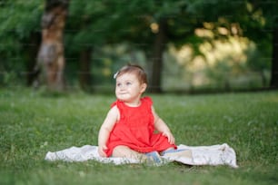 Cute adorable baby girl in red dress sitting on white blanket in park outdoors. Funny child toddler playing and having fun on summer day outside. Funny kid. Authentic lifestyle happy childhood.