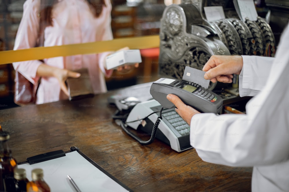 Unrecognizable woman customer making payment in ancient drugstore, bying the medicine. Pharmacist man holding terminal, and swiping patient's credit card credit card