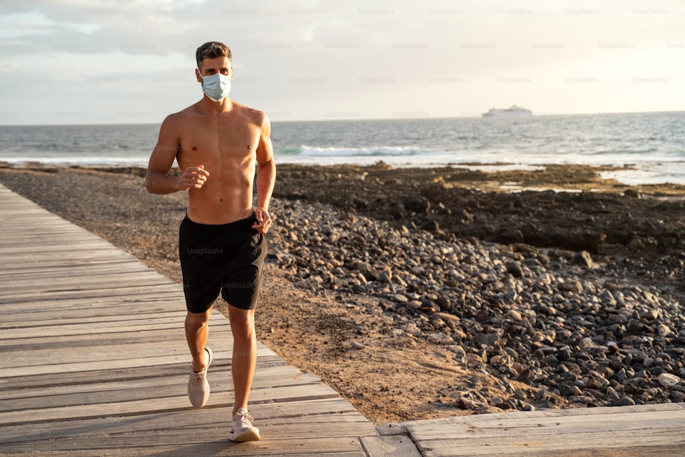 격리 기간 동안 아침에 바다 근처에서 운동하고 달리는 젊은 잘생긴 남자. 보호용 안면 마스크의 활동적인 건강한 라이프스타일 개념. 코로나 바이러스 COVID 19.