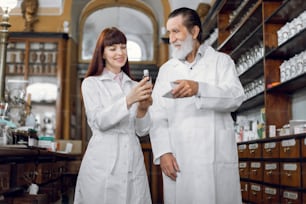 Dos farmacéuticos, un hombre mayor y una mujer joven que trabajan en una antigua farmacia. Farmacéuticos y farmacéuticas que comprueban la calidad de los medicamentos, comparando dos formas farmacéuticas de medicamentos.