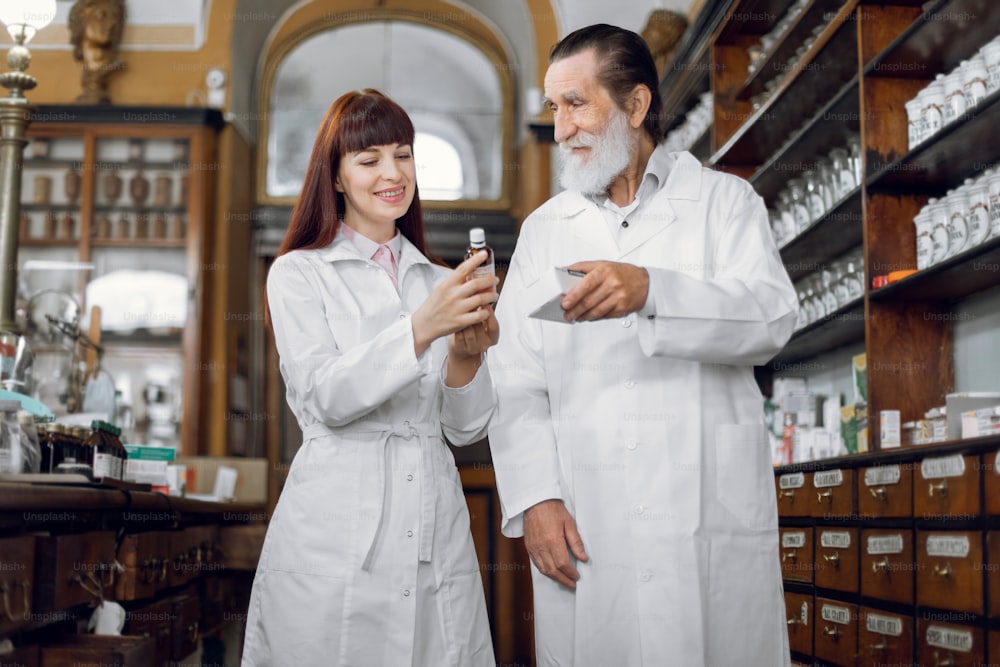 Two pharmacists, senior man and young woman working in vintage old drugstore. Male and female pharmacists checking medicines quality, comparing two dosage forms of medicine.