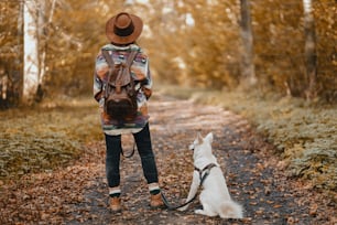Mujer elegante con mochila que camina con un adorable perro blanco en bosques soleados de otoño. Lindo cachorro de pastor suizo y hembra dueña que viaja en el bosque de otoño. Espacio para el texto