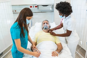 Doctors with protective masks are examining the infected aging patient in the hospital. In the Hospital Senior Patient Rests with oxygen mask, Lying on the Bed.