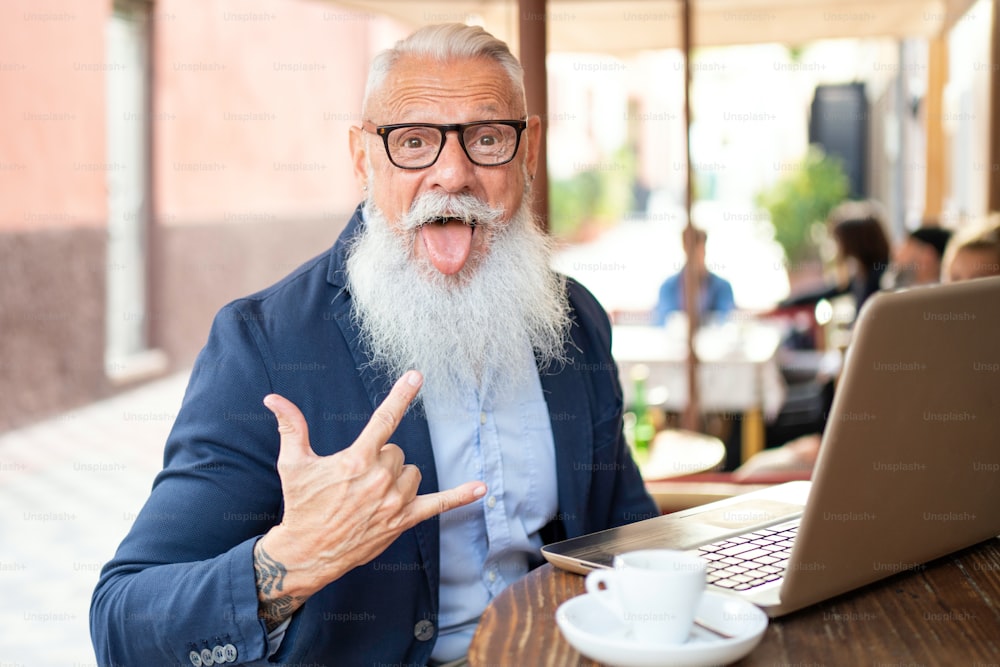 Smiling happy mature man with white stylish beard drinking coffee and using computer during lunch break at cafe outdoor. Laughing man using social media network technology feeling excited. Success and business lifestyle concept.