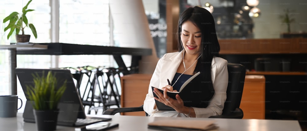 Happy businesswoman relaxing at her workplace listening music over earphones and making note on notebook.