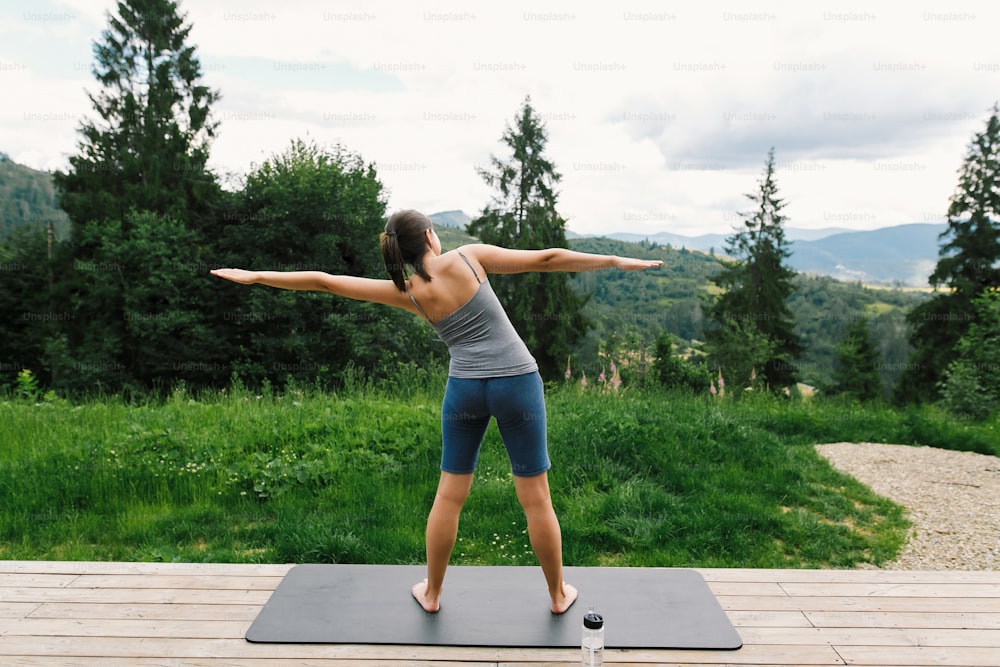 Mulher jovem que se estende no tapete de yoga no fundo das colinas ensolaradas das montanhas. Treino ao ar livre e treino. Fêmea esportiva casual no terraço de madeira entre as árvores, estilo de vida saudável
