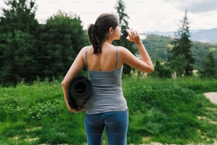 Giovane donna che beve acqua dopo l'allenamento sullo sfondo delle colline di montagna soleggiate. Allenamento all'aperto. Stile di vita sano. Femmina casual sportiva che tiene il tappetino da yoga e l'acqua potabile dalla bottiglia tra gli alberi