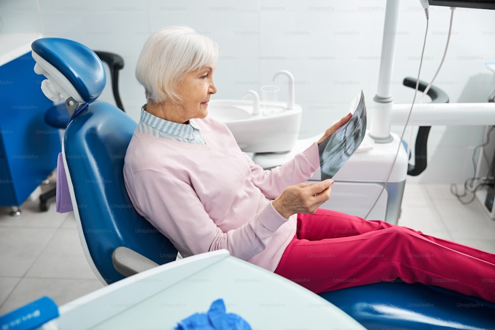 Woman on pension holding and checking radiograph of teeth while waiting on a dental chair