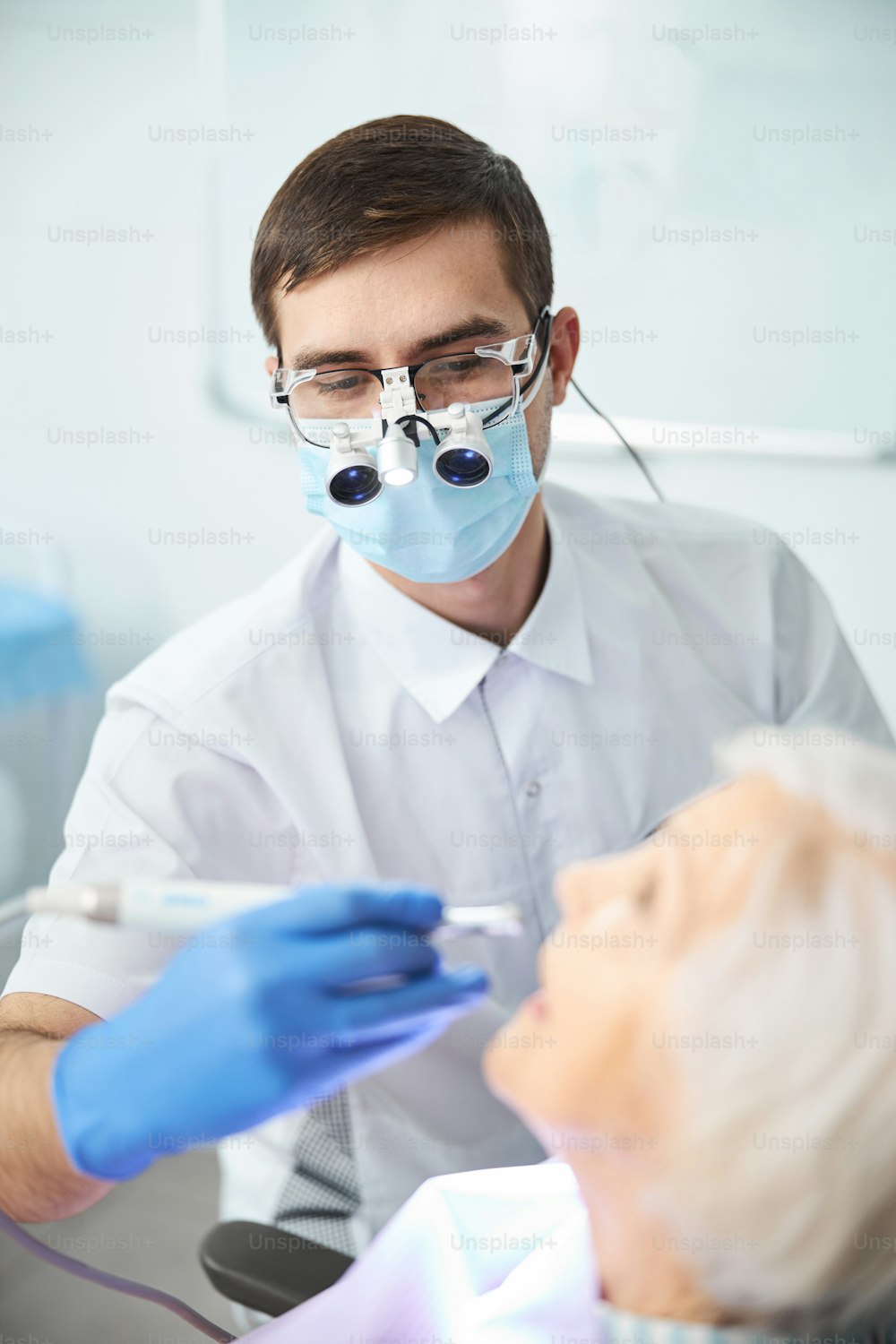 Dentista masculino en lupas binoculares con luz y máscara facial operando una pieza de mano durante un procedimiento en los dientes de la mujer