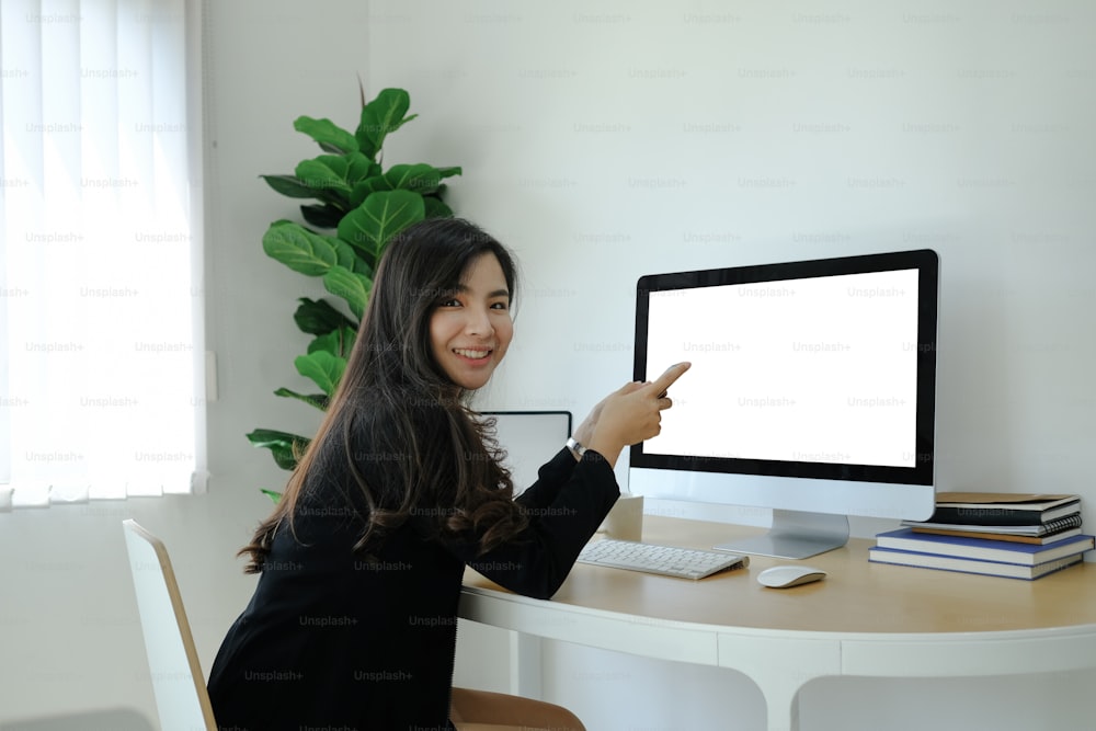 Side view of happy businesswoman holding mobile phone and smiling to camera while sitting in front of computer at workplace.