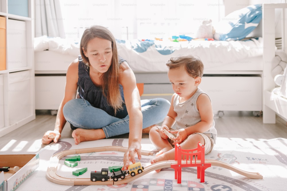 自宅の床で木造の鉄道で遊んでいる母親と幼児の男の子。幼児教育の発達。線路を建設したり、教育用のおもちゃの電車で遊んだりする子供たち。子供のための余暇活動。