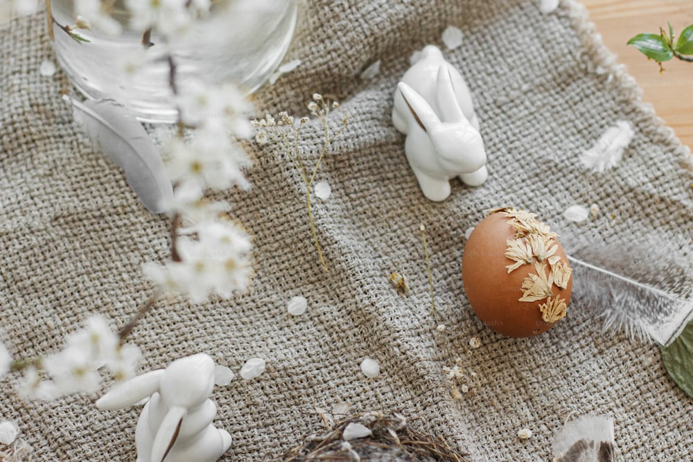 Osterei verziert mit trockenen Blütenblättern und Hase auf rustikalem Tisch mit Leinenserviette, Federn und Kirschblüten. Kreatives natürliches, umweltfreundliches Dekor von Ostereiern. Frohe Ostern