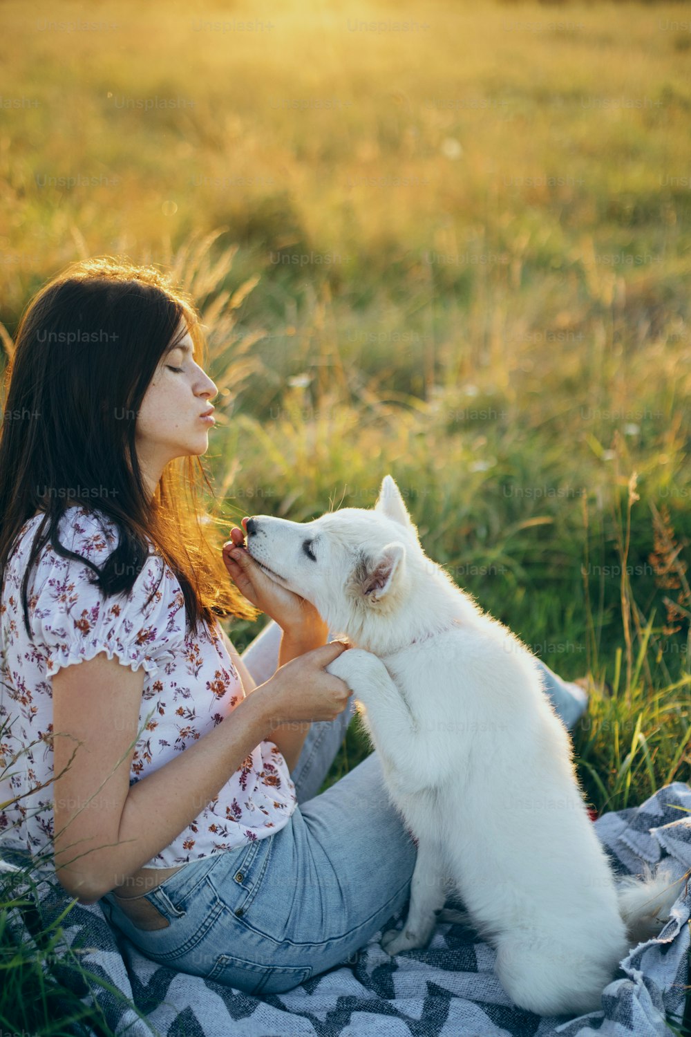 Mujer feliz entrenando a un lindo cachorro blanco para que se comporte en el prado de verano a la luz del atardecer. Elegante mujer joven casual que se relaja con un adorable cachorro esponjoso de pastor suizo. Vacaciones con mascota