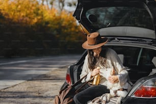 Stilvolle junge Frau sitzt mit süßem weißen Hund im Kofferraum an der sonnigen Herbststraße. Roadtrip mit Haustier. Reisen mit einem Schweizer Schäferhundwelpen im Land aufgrund der Coronavirus-Pandemie. Platz für Text