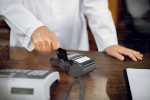 Cashless payment at pharmacy. Cropped close up shot of hands of male unrecognizable pharmacist in lab coat, working at pharmacy and swiping credit card through the terminal.