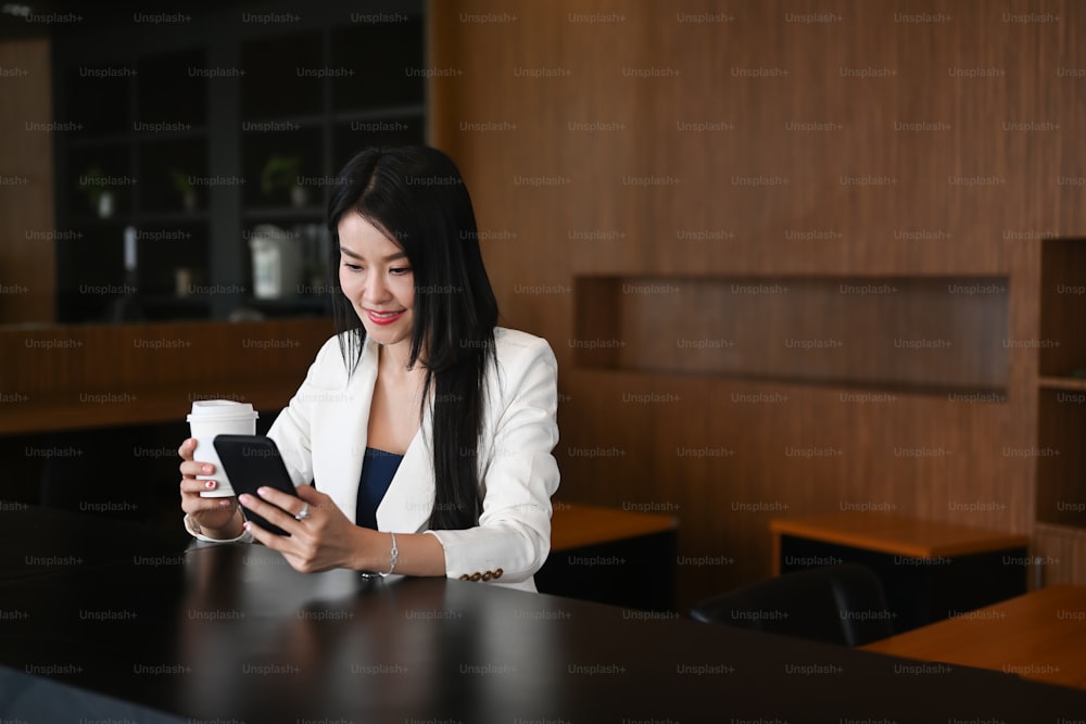 Smiling businesswoman reading news on smart phone and drinking hot coffee at office.