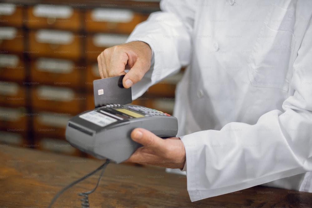 Bargeldloses Bezahlen in der Apotheke. Nahaufnahme von Händen eines männlichen, nicht erkennbaren Apothekers im Laborkittel, der in der Apotheke arbeitet und die Kreditkarte durch das Terminal zieht.