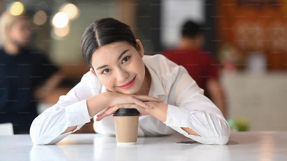 Cheerful young female office worker sitting with coffee cup and smiling to camera.
