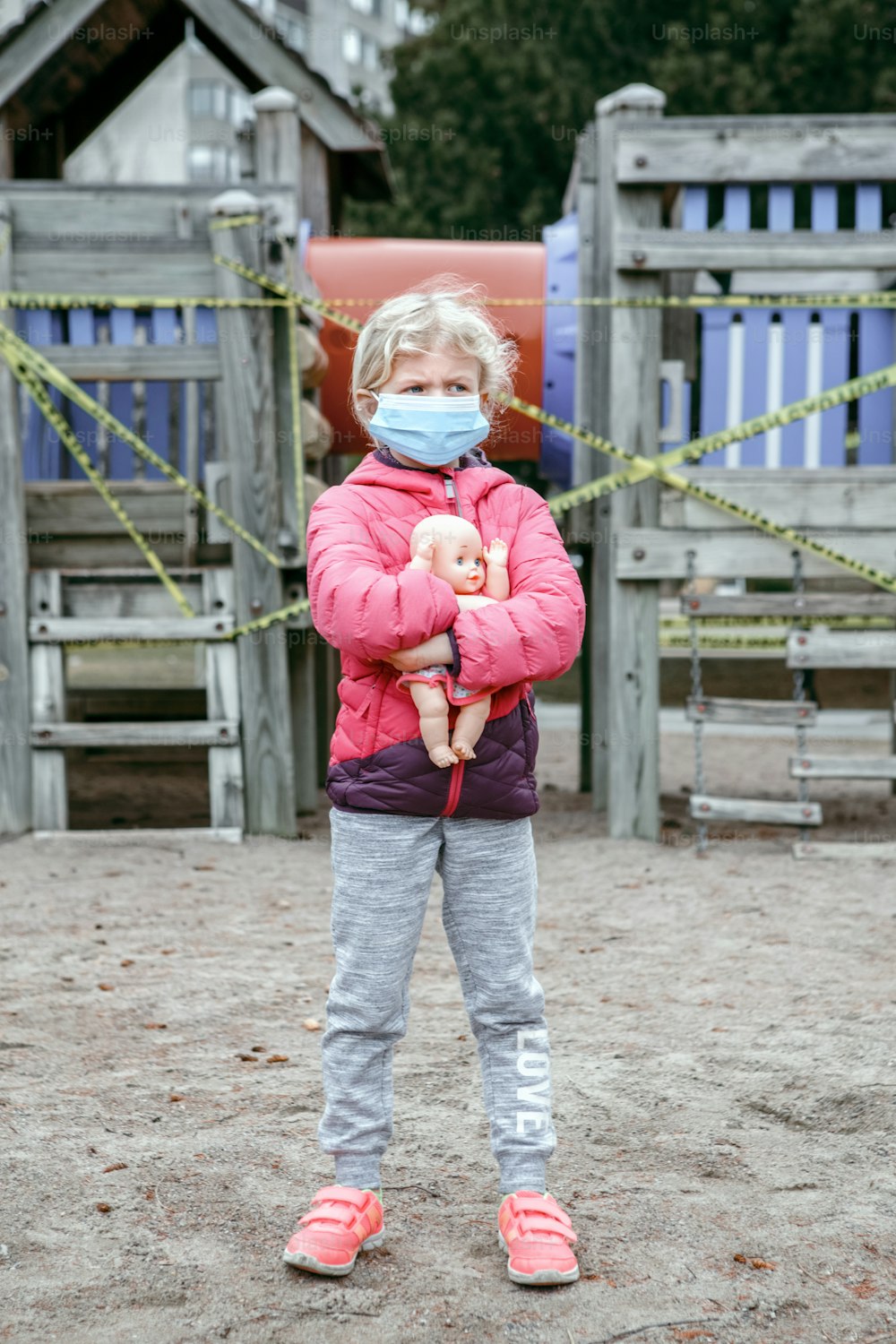 닫힌 놀이터 야외에서 아기 장난감을 가진 얼굴 마스크를 쓴 슬픈 백인 소녀. 캐나다 토론토에서 노란색 주의 테이프로 잠긴 어린이 놀이 공간. 코로나 바이러스 사회적 거리 격리.