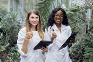 Equipo de dos mujeres multiétnicas jóvenes sonrientes, agrónomos científicos profesionales, posando ante la cámara con una sonrisa y pulgares hacia arriba, mientras están de pie en un hermoso invernadero con plantas tropicales.