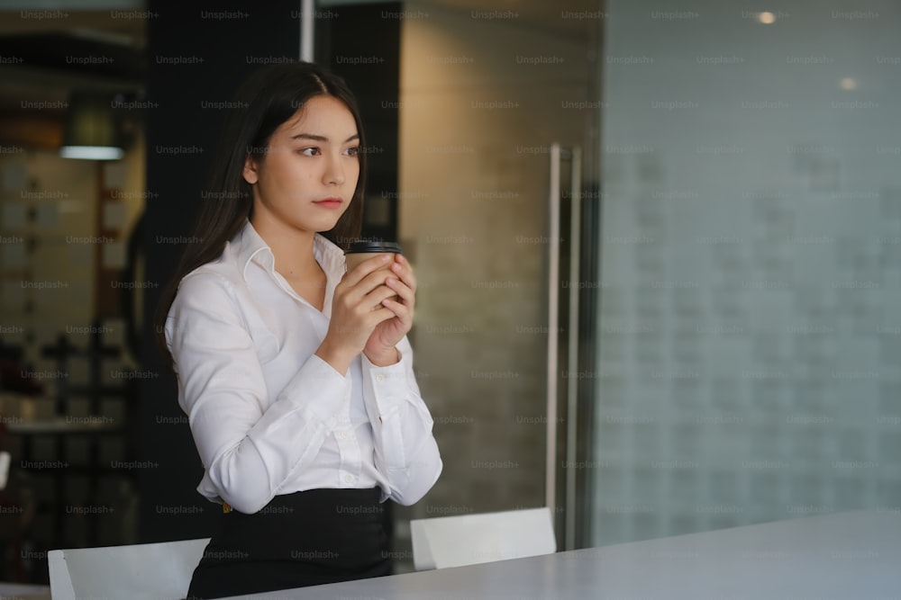 Young woman office worker holding coffee cup or tea and looking away thoughtfully.