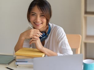 Ritratto di bella femmina che sorride alla macchina fotografica mentre è seduta sul posto di lavoro con laptop e pila di libri